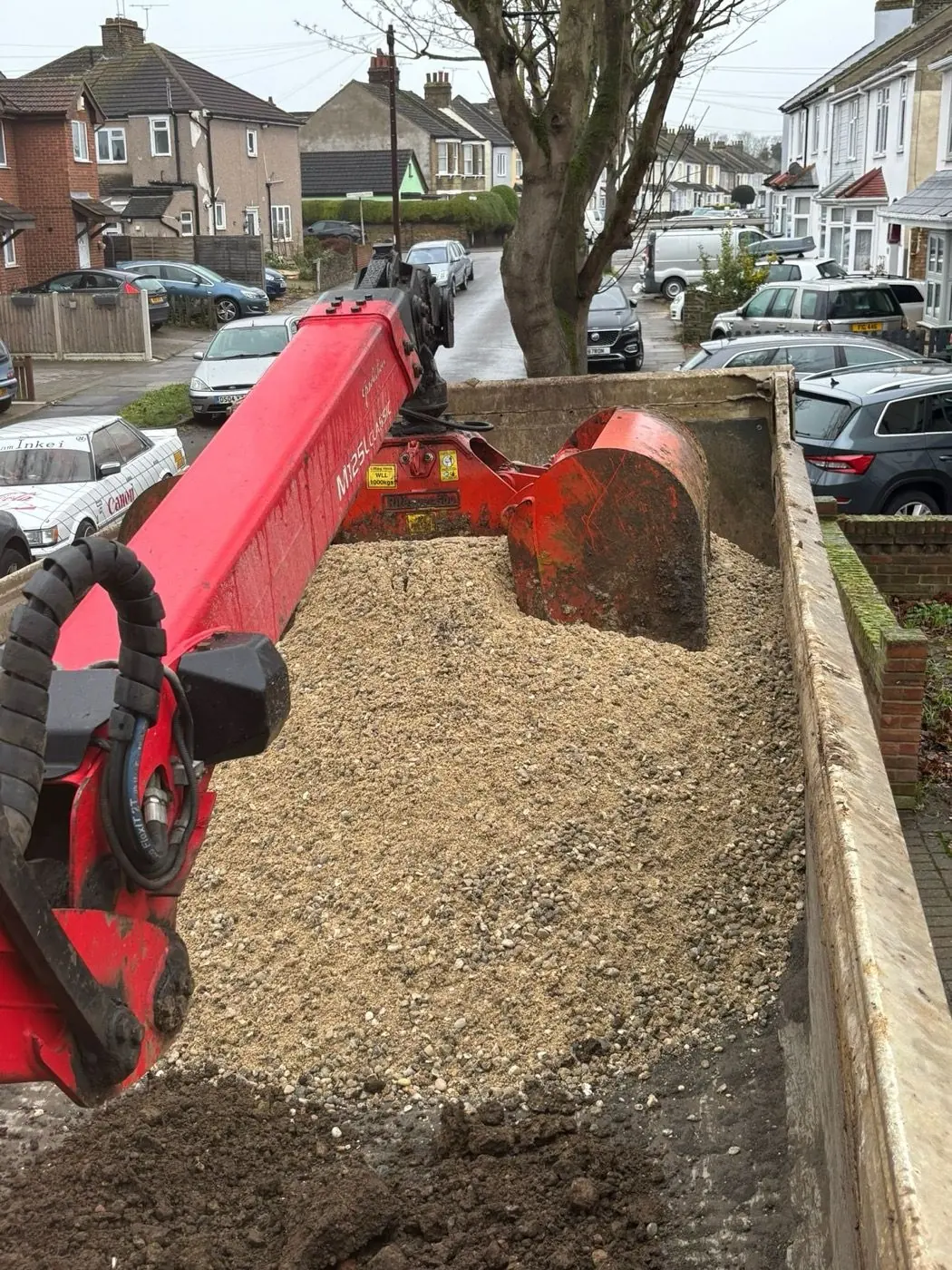 Grab bucket above a lorry bed containing light-coloured loose aggregate